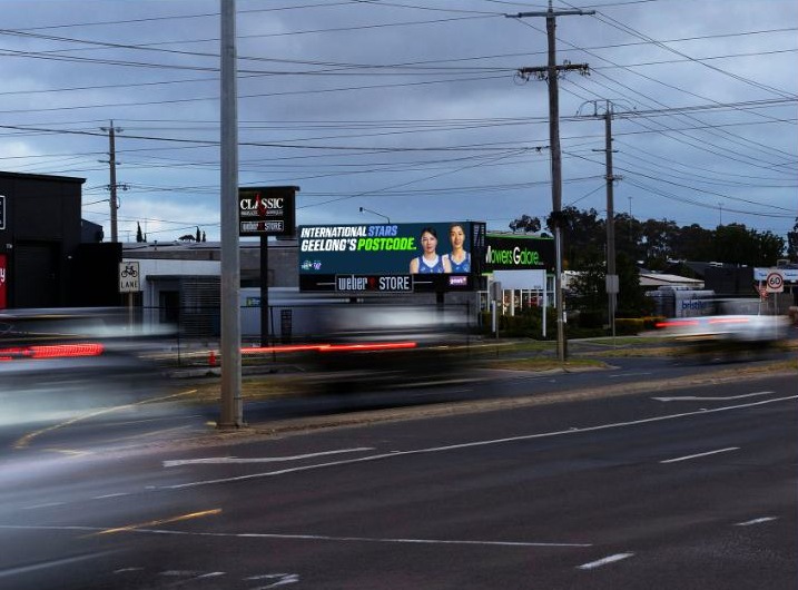 Illuminated digital billboard advertising Geelong basketball beside a busy road at dusk